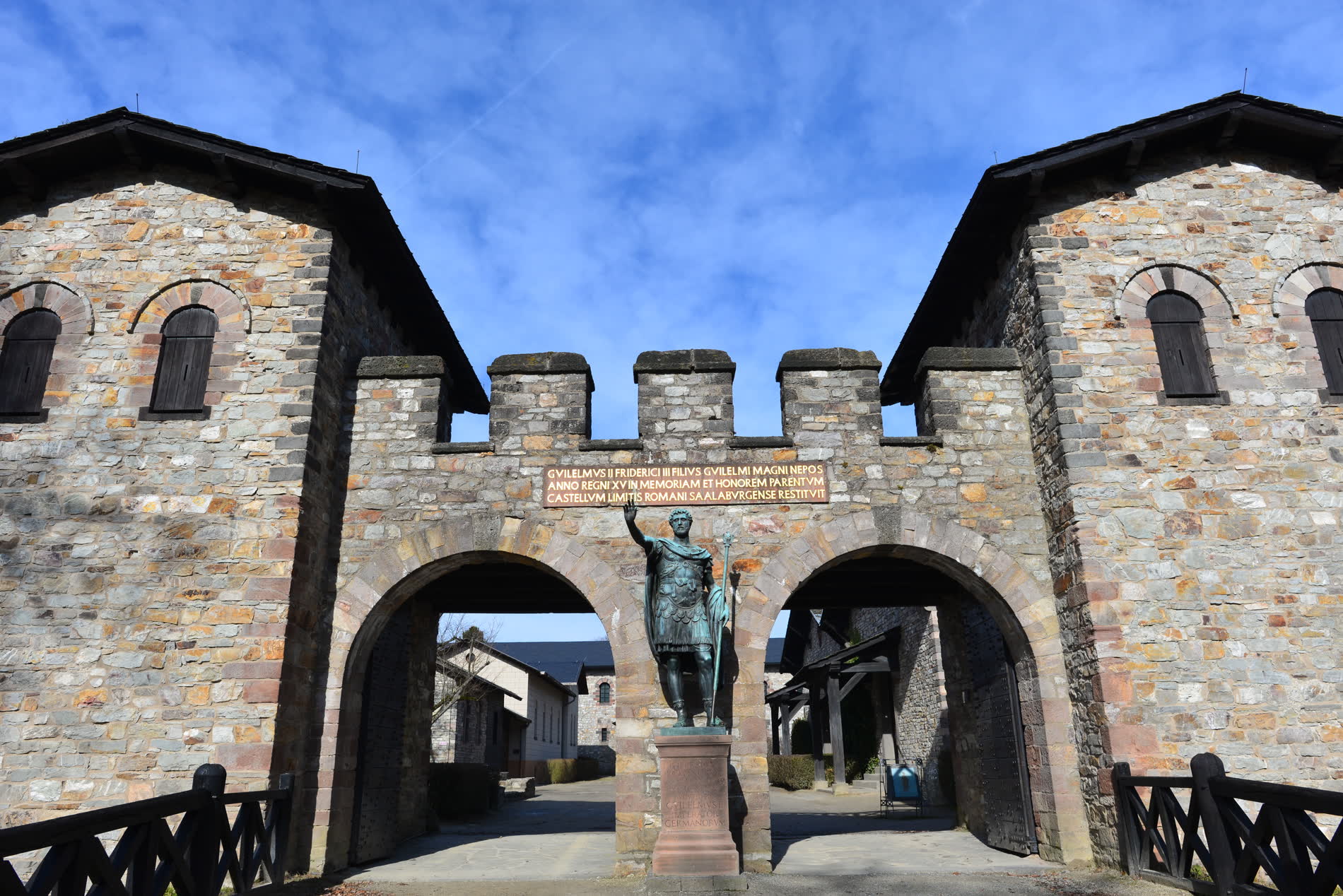 Steinernes Tor mit zwei Durchfahrten und Zinnen, davor eine Bronzestatue eines römischen Kaisers Steinernes Tor mit zwei Durchfahrten und Zinnen, davor eine Bronzestatue eines römischen Kaisers