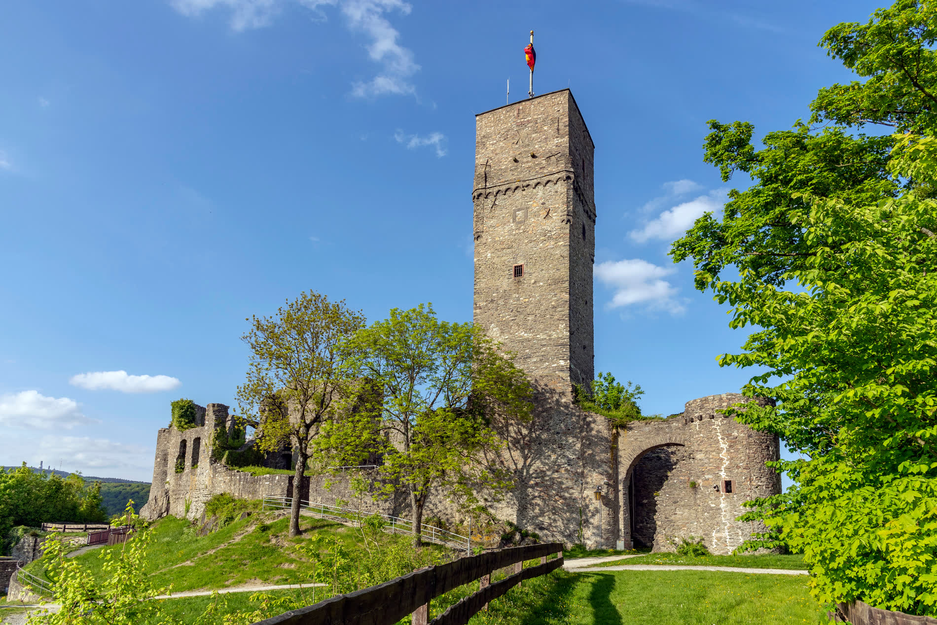 Hoher quadratischer Bergfried mit Fahne, daneben Mauerreste und ein Rundturm mit Torbogen Hoher quadratischer Bergfried mit Fahne, daneben Mauerreste und ein Rundturm mit Torbogen